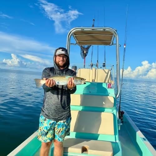 Man in fishing attire holding a freshly caught fish on a small boat in calm blue waters under a partly cloudy sky.