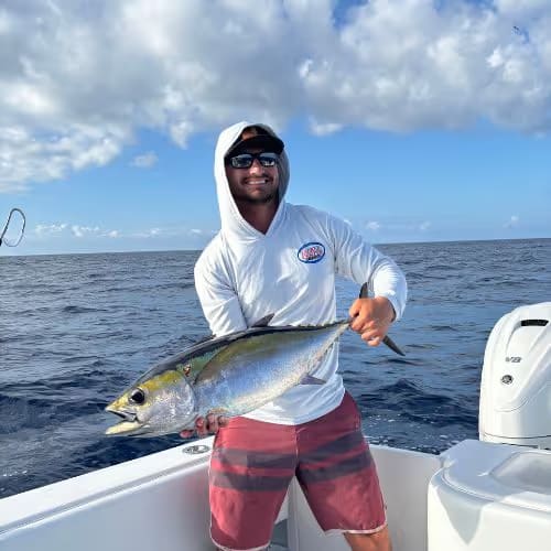Man wearing sunglasses and a hooded shirt holding a large tuna on a boat in the ocean.