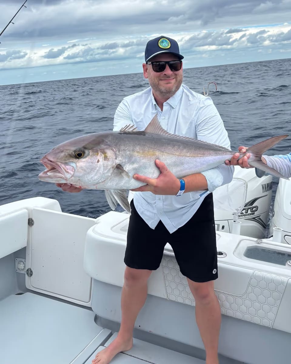 Man wearing sunglasses and a cap holding a large fish on a boat with ocean and cloudy sky in the background.
