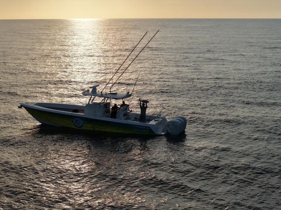 A Fishing Guru boat on calm ocean water at sunset with people on board, one holding up a large fish.