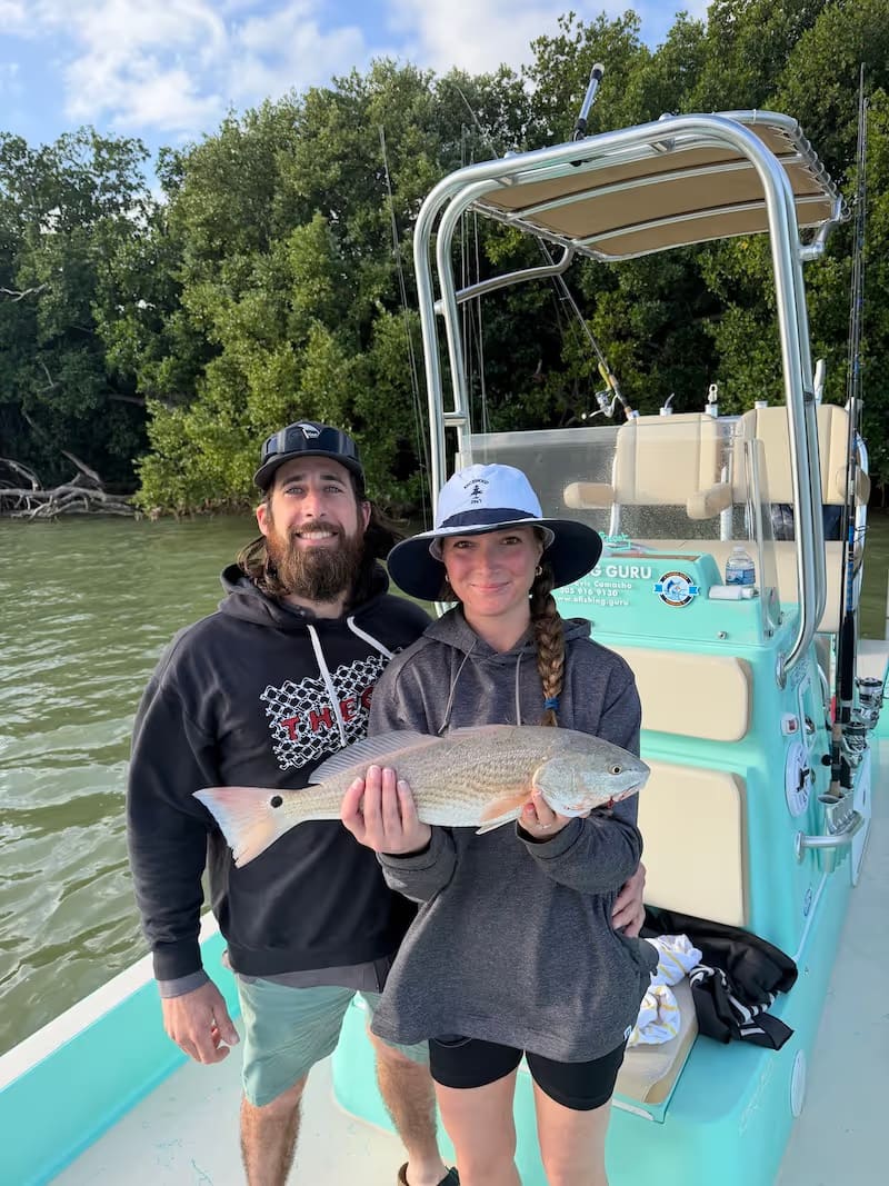 Man and woman on a boat near mangroves holding a large redfish, smiling at the camera.