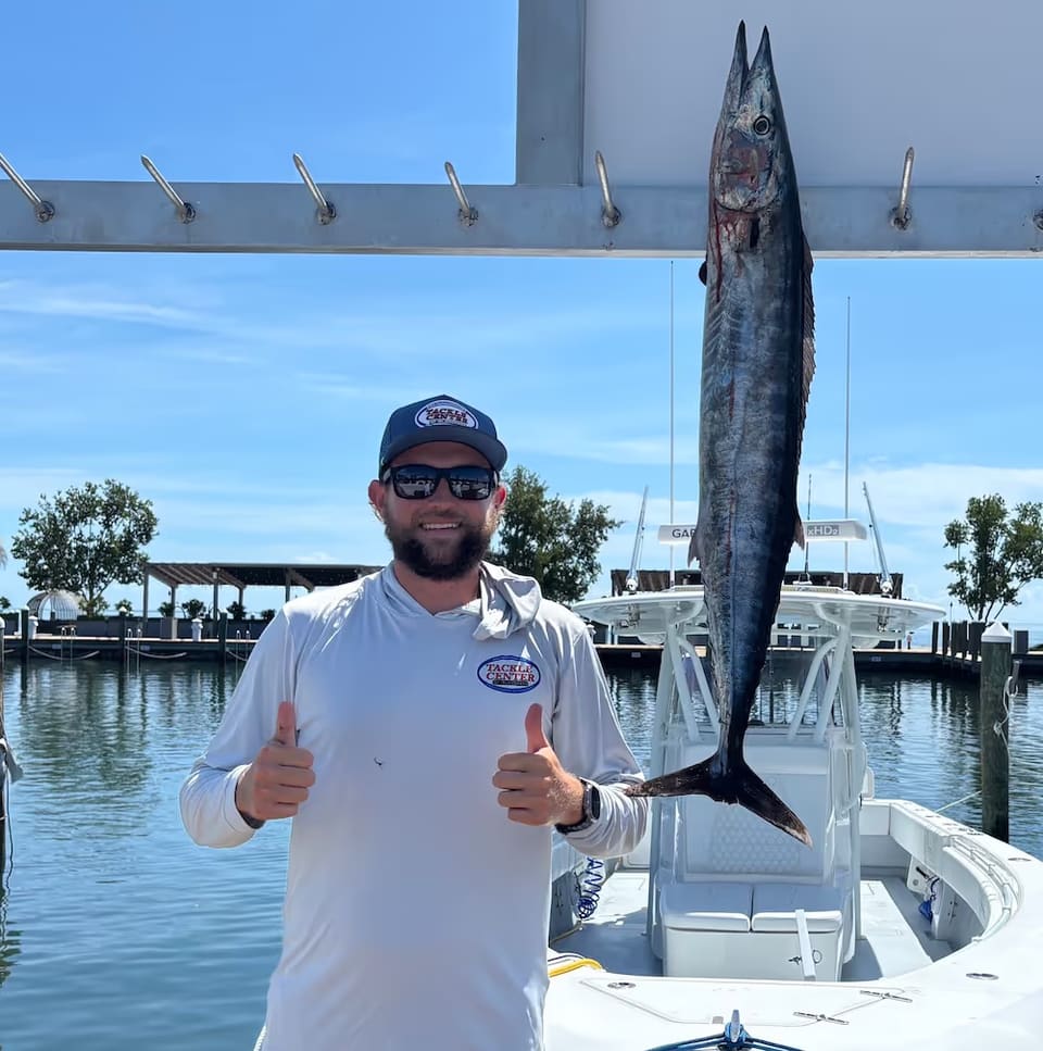 Man wearing sunglasses and a white shirt giving two thumbs up next to a large fish hanging on a boat dock.