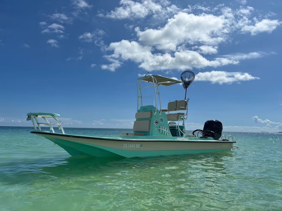 A mint green and white flats fishing boat floating on clear green water under a blue sky with scattered clouds.