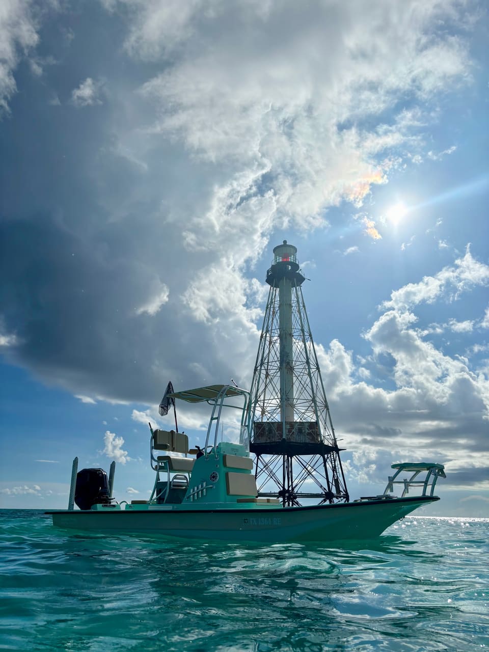 Small boat floating near a tall metal lighthouse under a partly cloudy sky with the sun shining.
