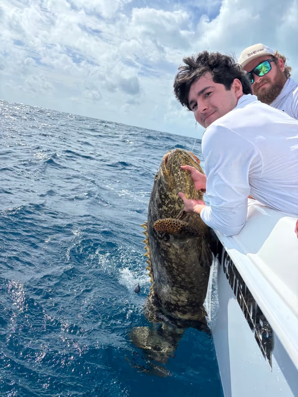 Two men on a boat holding a large grouper fish partially in the water under a cloudy sky.