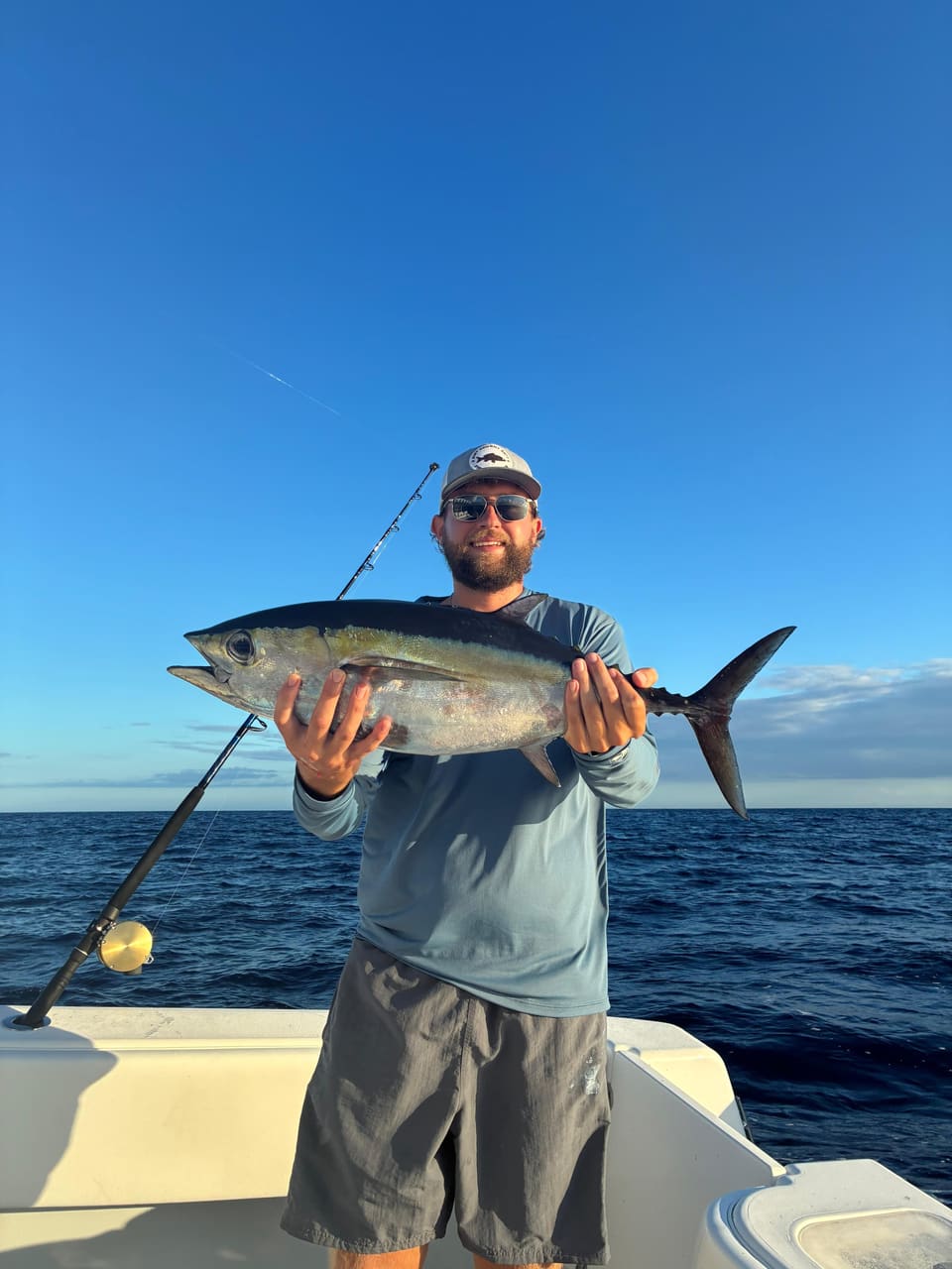 Man wearing sunglasses and a cap holding a large fish on a boat with ocean and clear blue sky background.