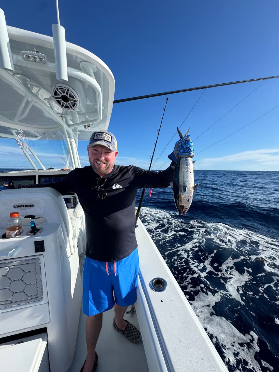 Angler holding up a blackfin tuna on the stern of a charter boat in the open ocean, with deep blue water and a clear blue sky behind him