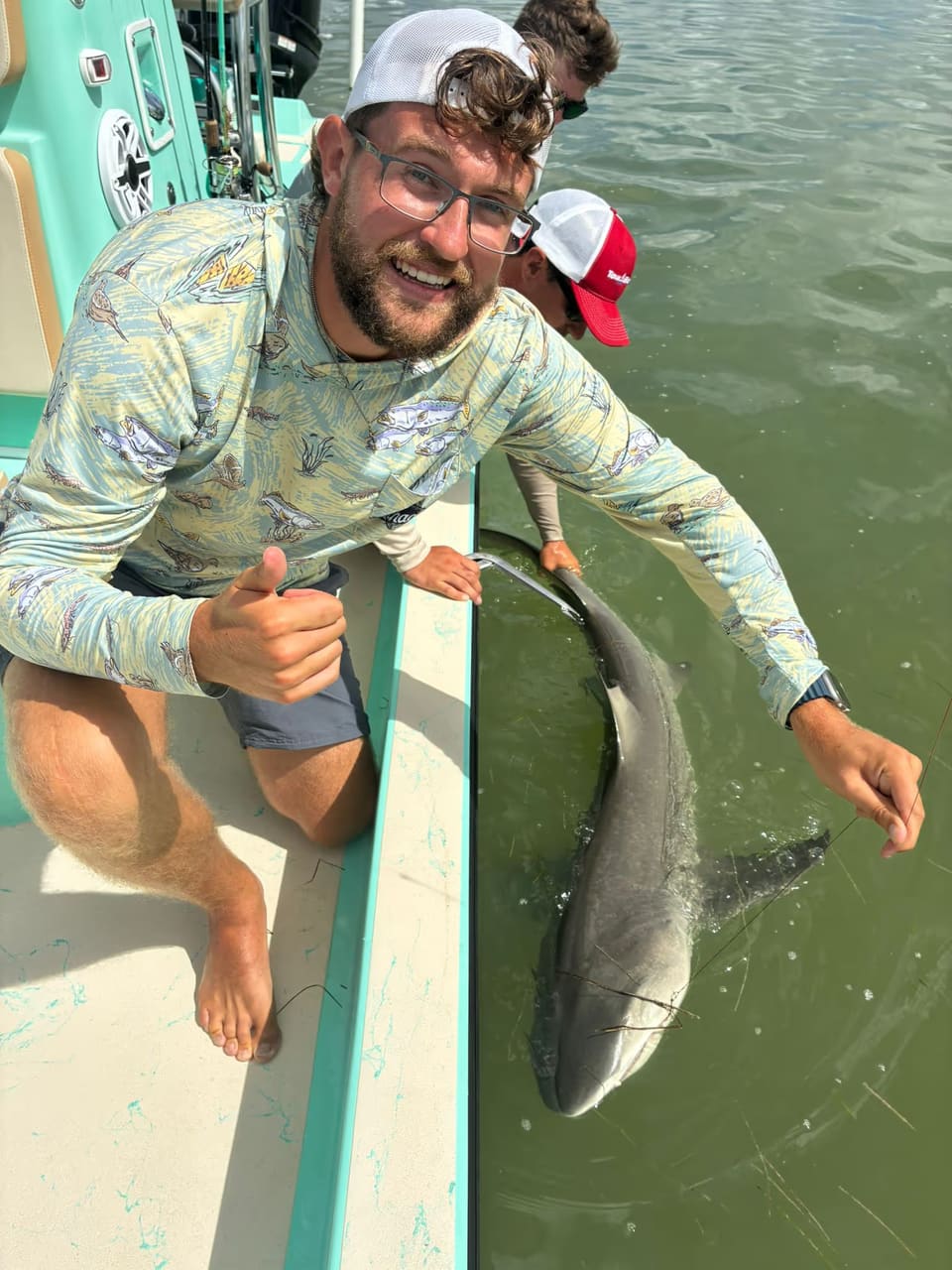 Smiling man giving thumbs up on a boat while holding a shark in the water.