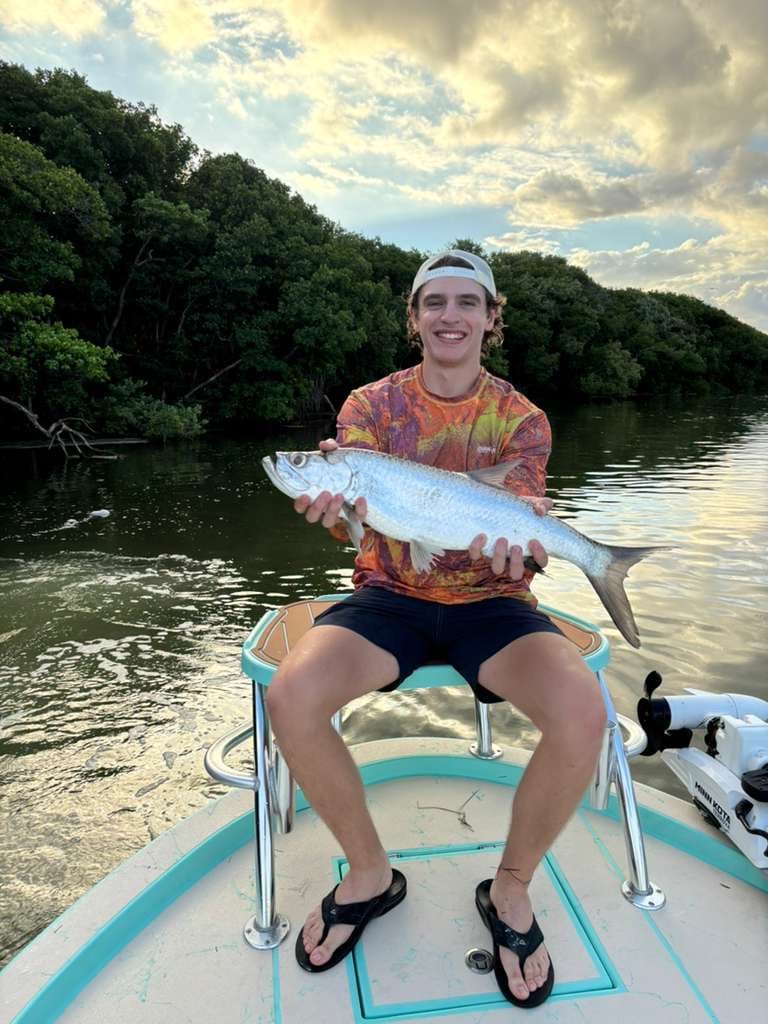 Smiling young man sitting on a boat holding a large silver fish with water and trees in the background.