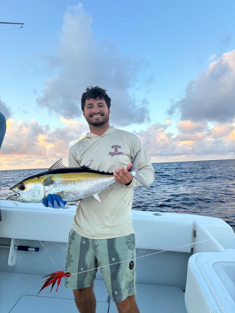 Smiling man on a boat holding a freshly caught yellowfin tuna with the ocean and cloudy sky in the background.