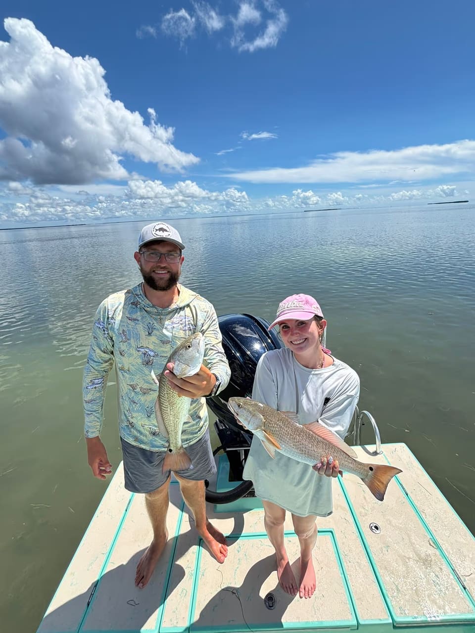 A couple standing on the bow platform of a flats boat, each holding a redfish they caught, with calm shallow water and puffy clouds behind them