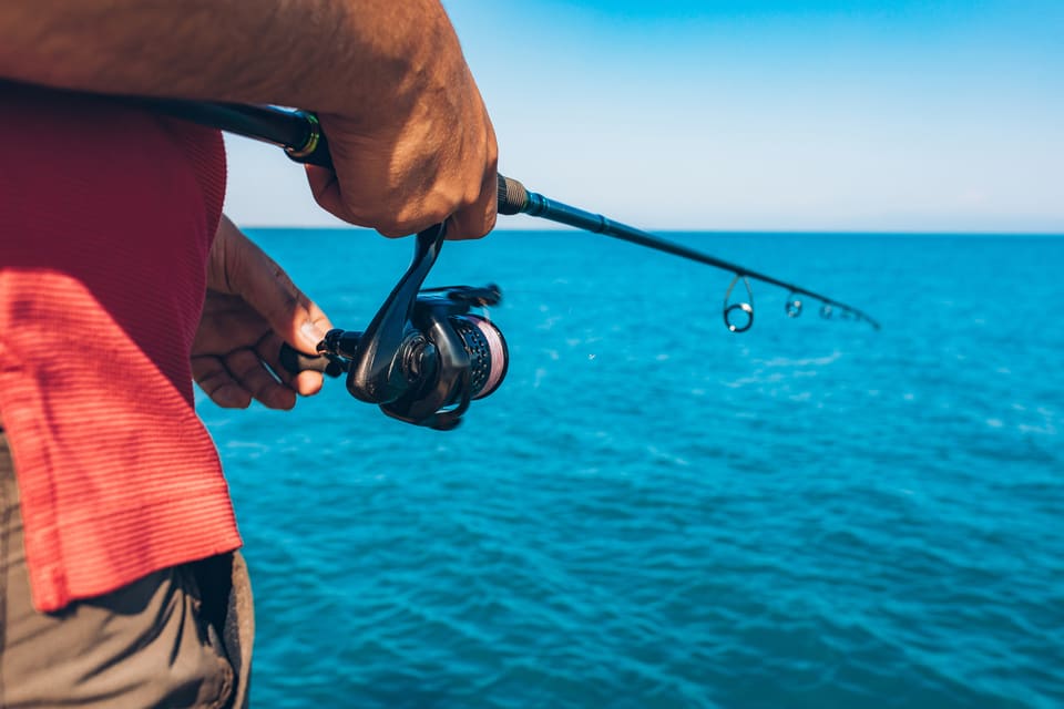Close-up of an angler's hands holding a spinning fishing reel and rod over brilliant blue ocean water on a clear day