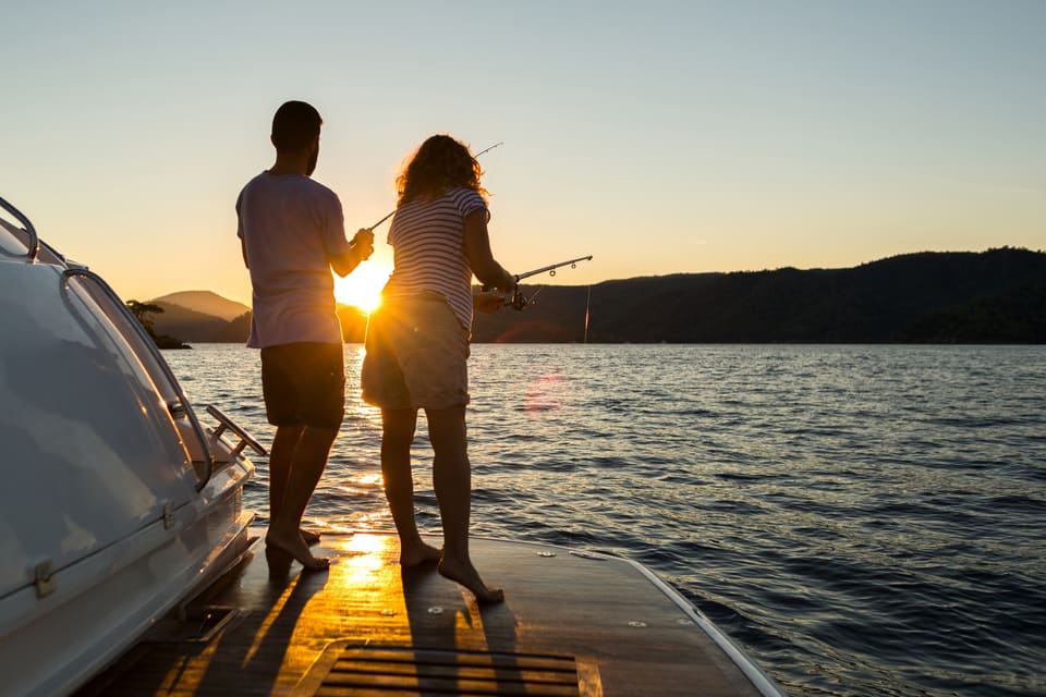 Silhouettes of a couple fishing together from the bow of a boat at sunset, with golden light reflecting on calm water and forested hills in the background