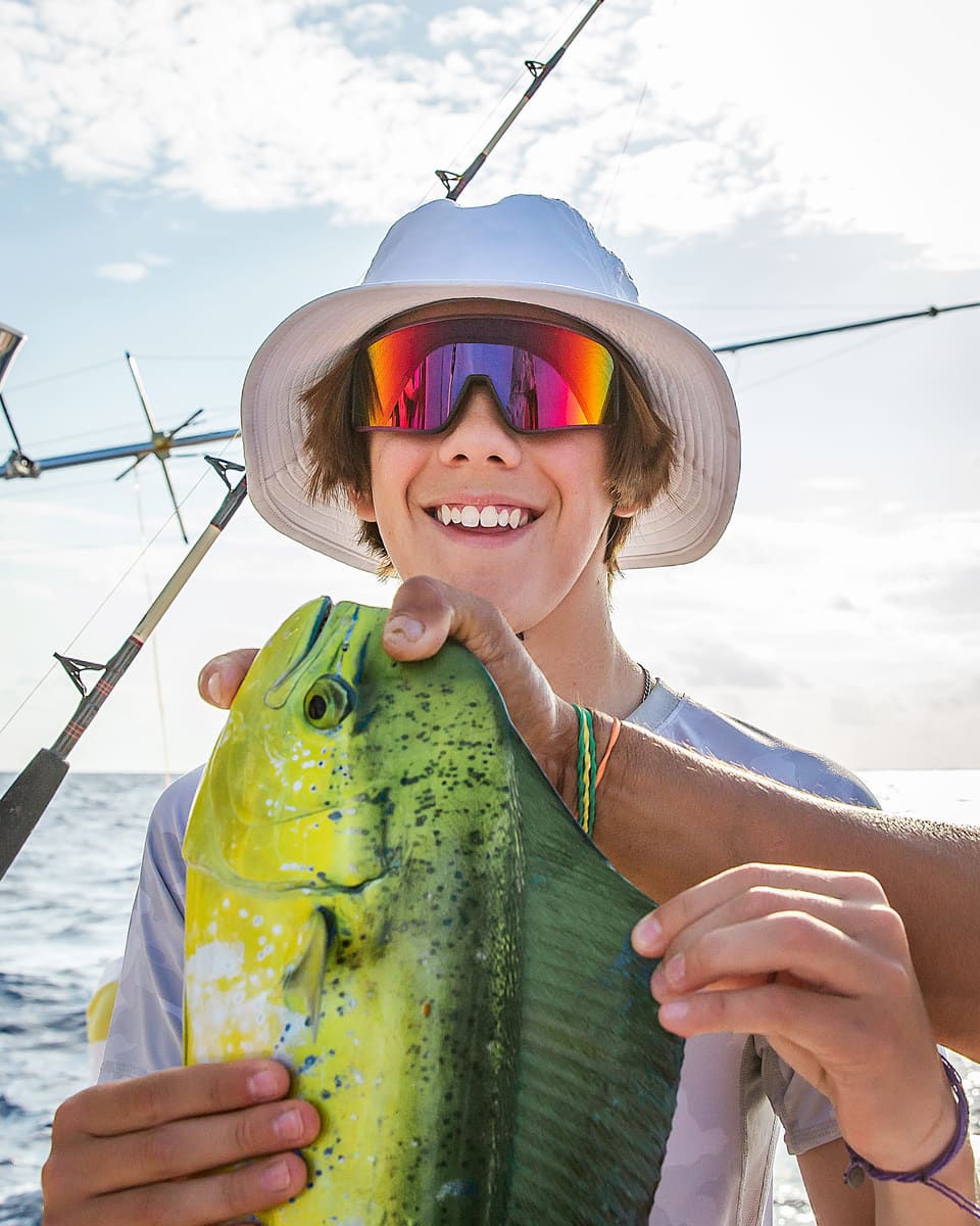 Teenage boy in a white bucket hat with colorful sunglasses holding a green and yellow Mahi Mahi caught off the coast of Islamadora.