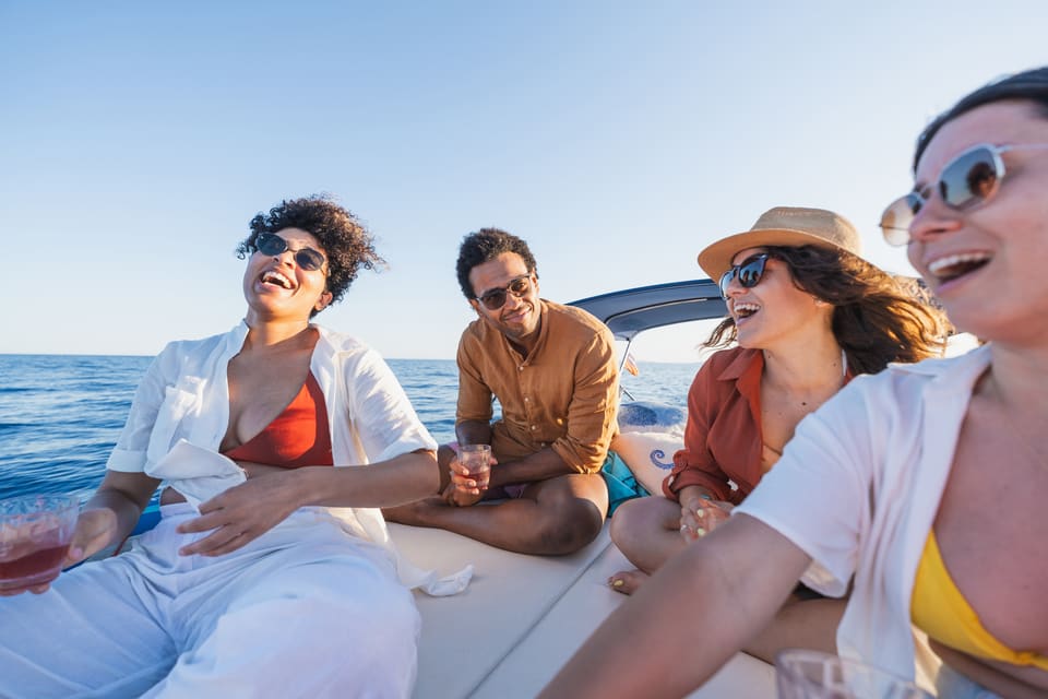 Four friends laughing together on a boat in bright sunlight on the open ocean, wearing sunglasses and summer clothing