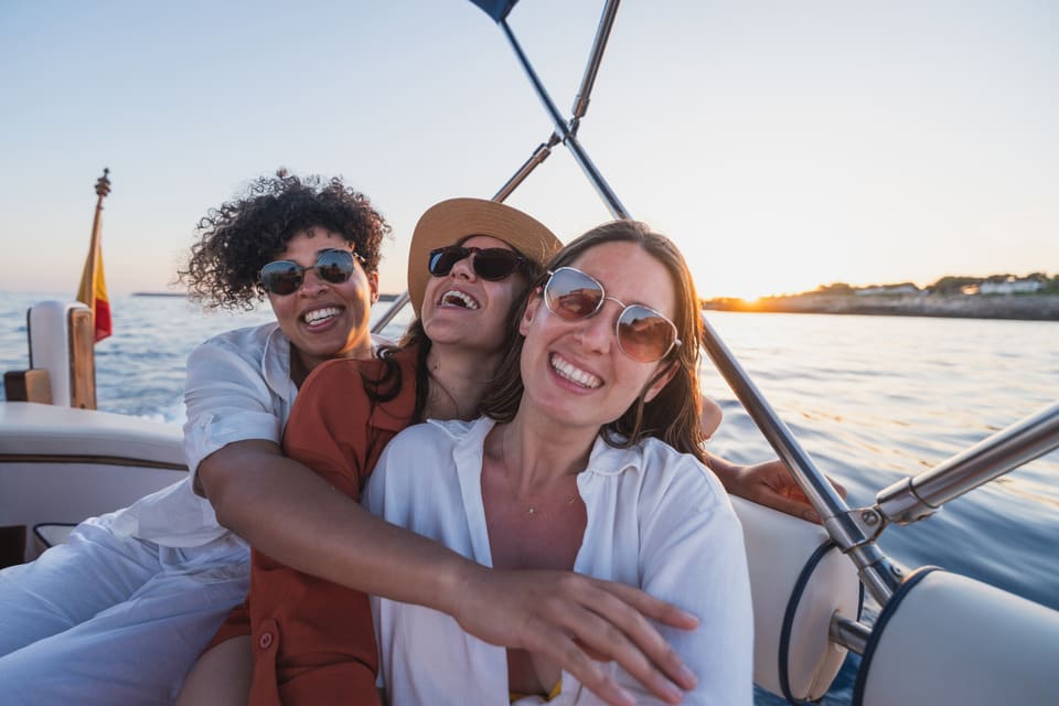 Three women laughing and embracing on a boat at golden hour, with calm ocean water and a glowing coastline behind them