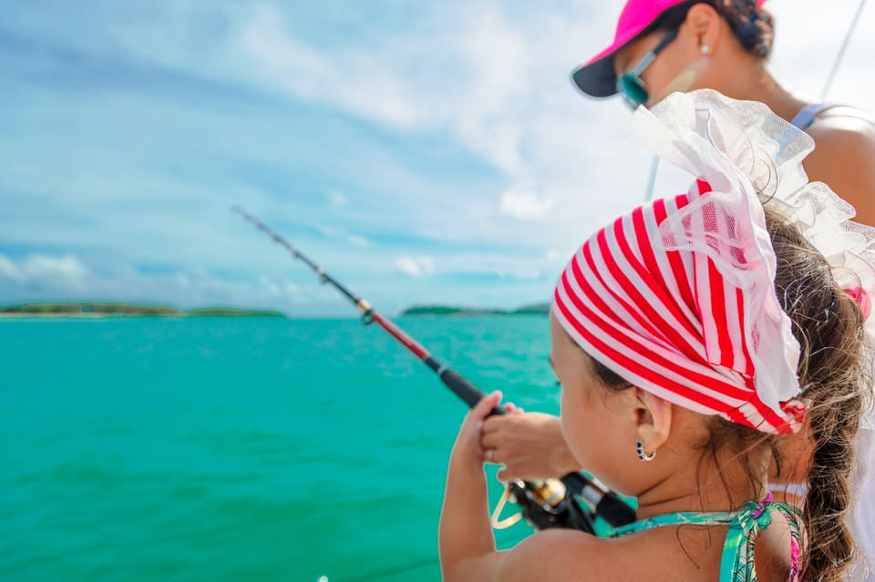 A young girl in a red-striped bandana holds a fishing rod over turquoise water on a boat, guided by her mother in a pink cap standing behind her
