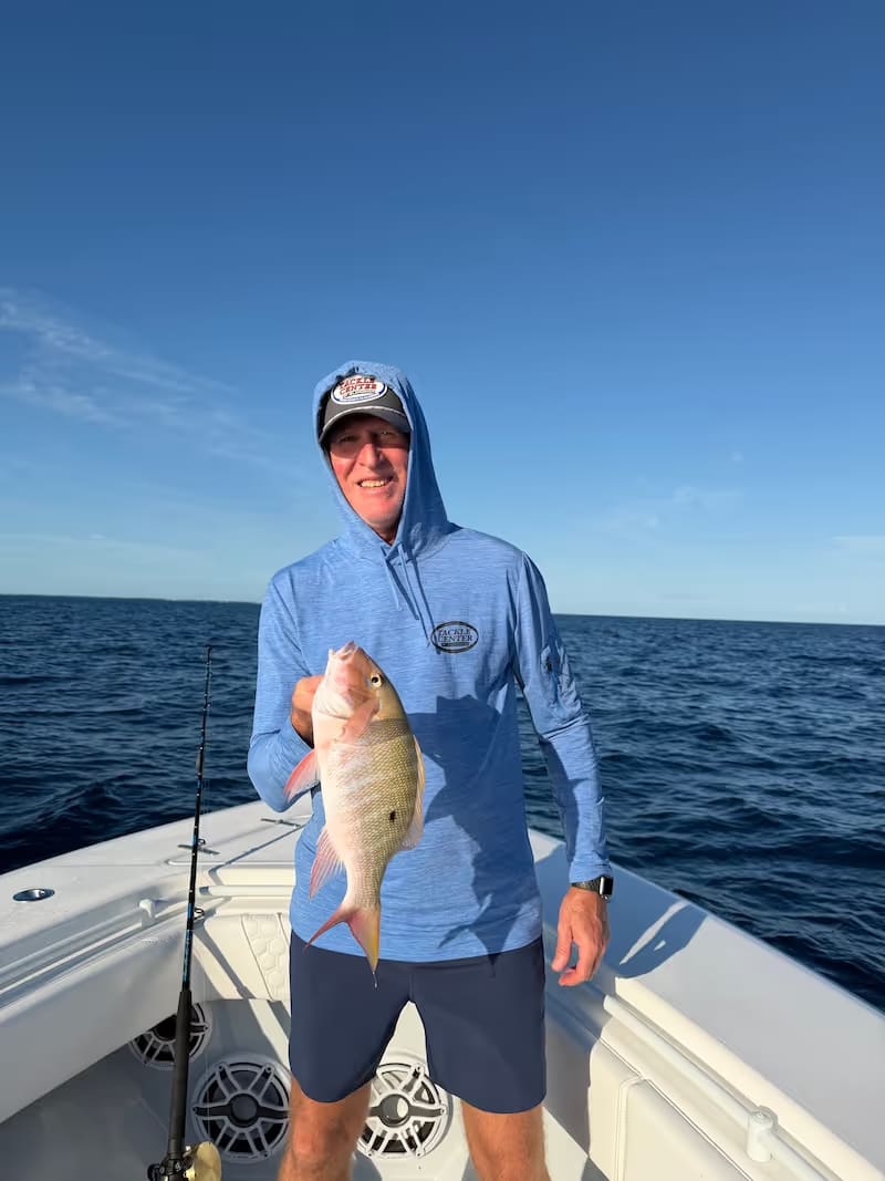Man in a blue hoodie and hat holding a mutton snapper on a boat with the ocean and clear sky in the background.