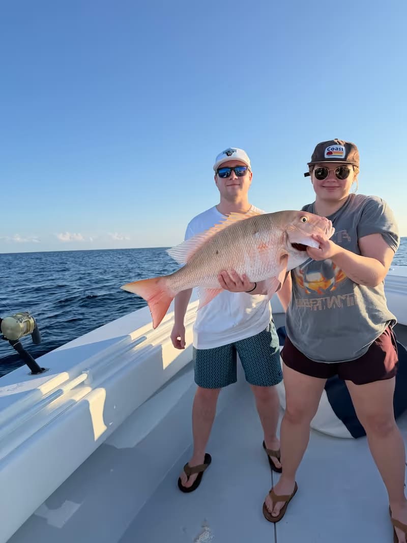 Two people on a boat holding a large pink snapper fish against a backdrop of the ocean and clear sky.
