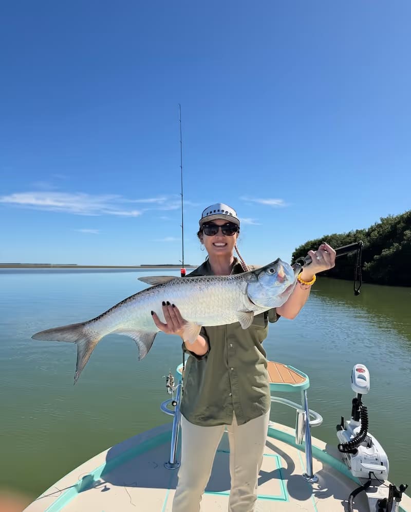 Woman wearing sunglasses and cap holding a large tarpon on a boat in calm water under a clear blue sky.