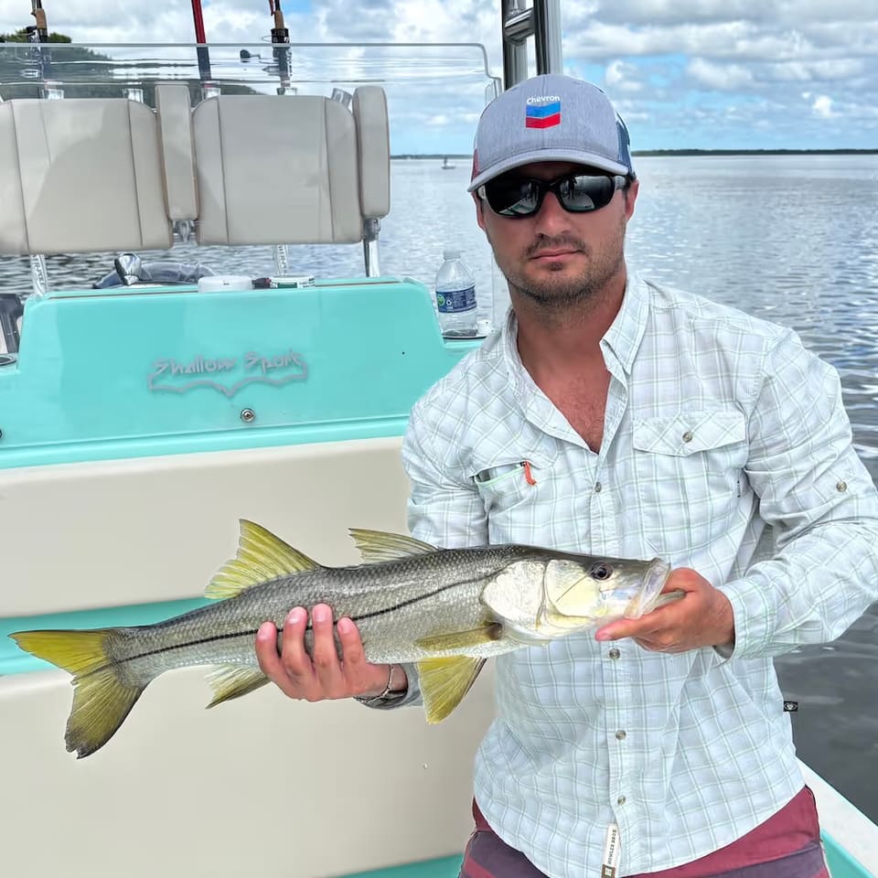 Man wearing sunglasses and a Chevron cap holding a snook on a boat with water and cloudy sky in the background.