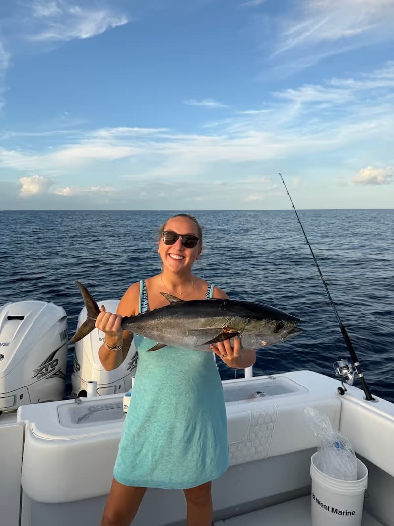 Smiling woman wearing sunglasses holding a large tuna on a boat in the open sea under a partly cloudy sky.